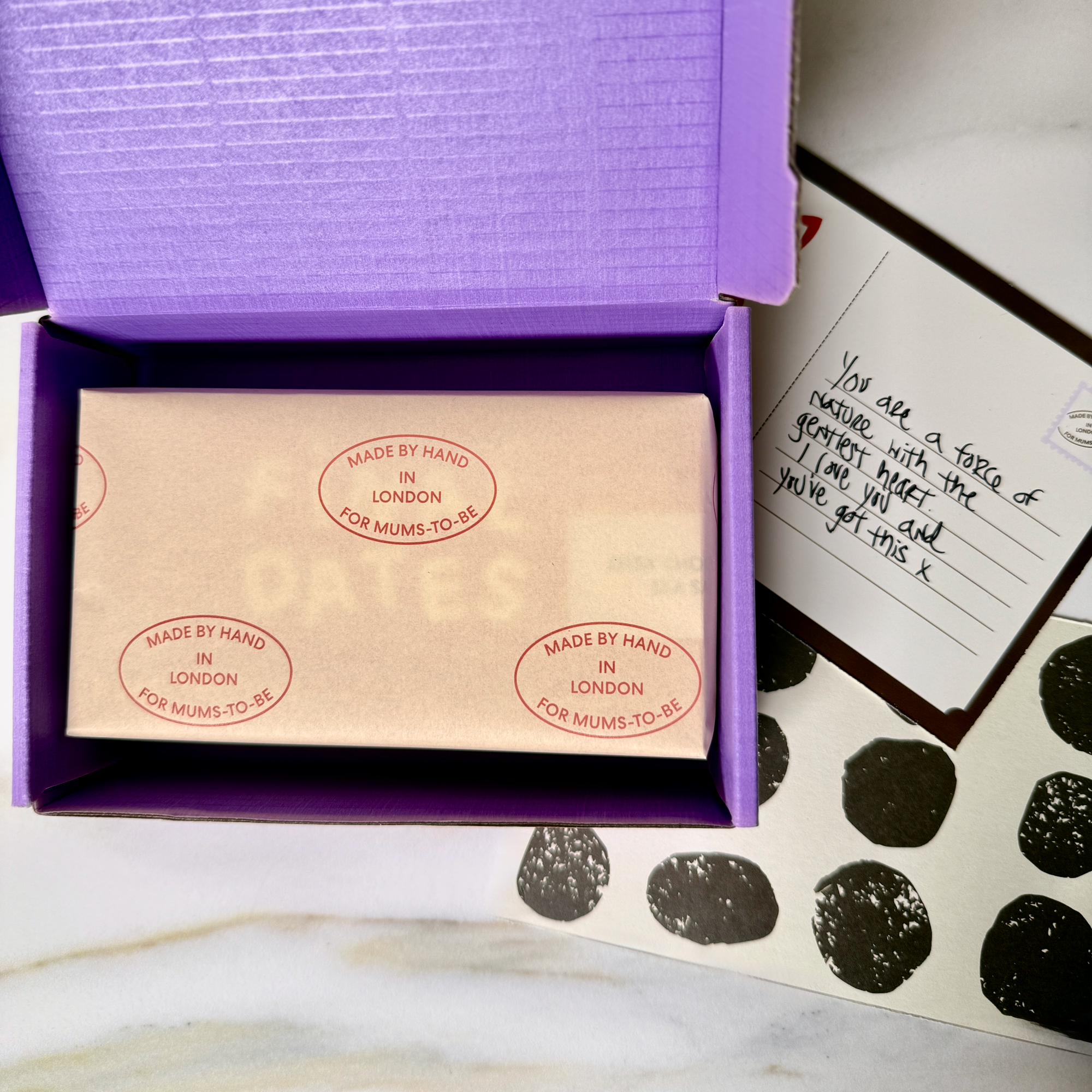 A purple gift box is open on a marble surface, revealing a gift wrapped box of truffles. A handwritten note and black-and-white polka dot card are placed beside it.